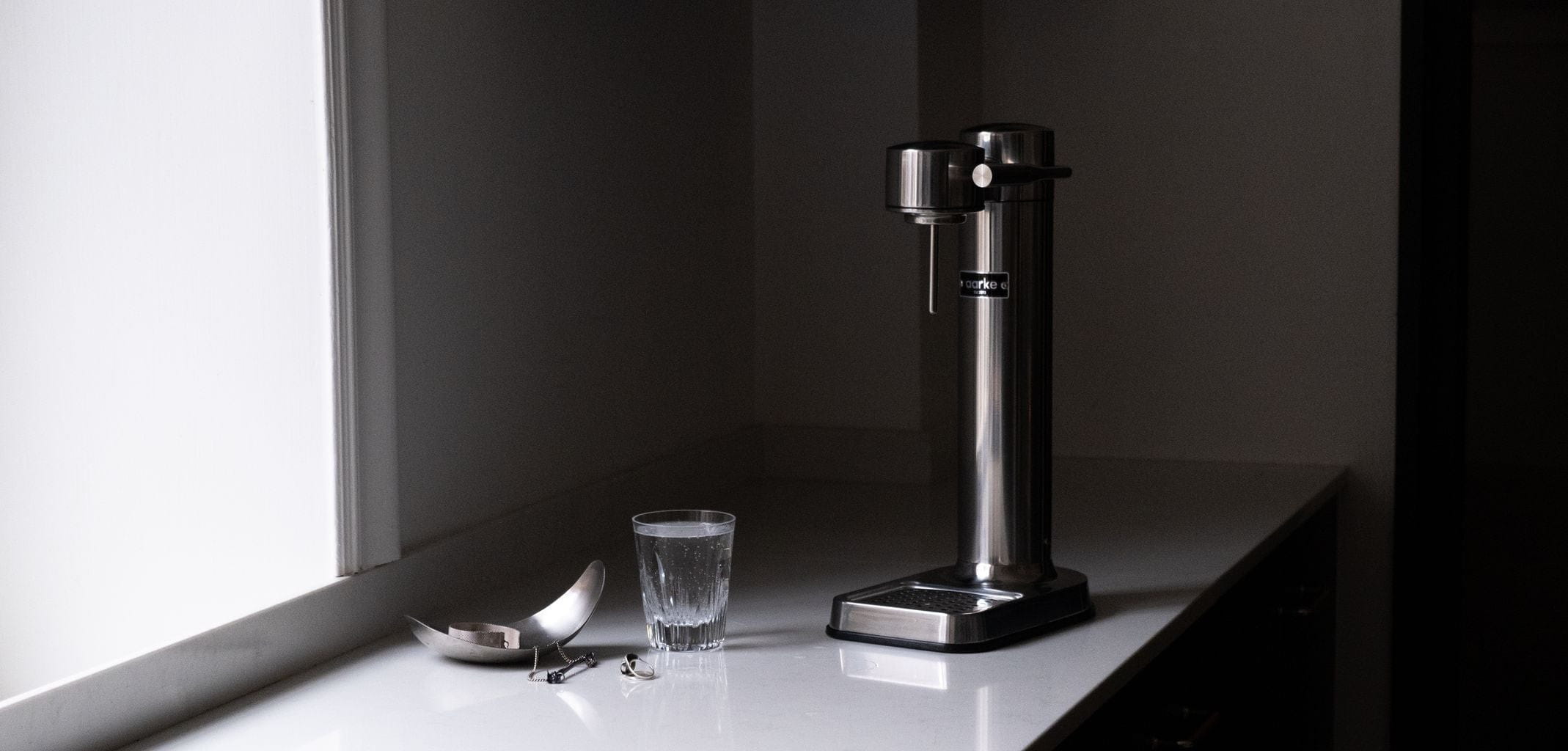 An Aarke Carbonator 3 Soda Maker, Polished Steel sits on a table next to a window.
